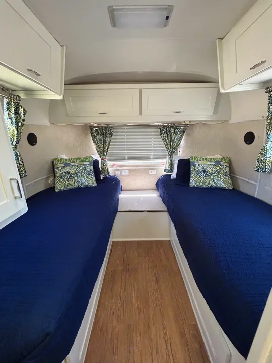 Compact RV rental bedroom interior with two twin beds featuring dark blue bedding, overhead cabinets, and patterned curtains.. Airstream Excella 2000