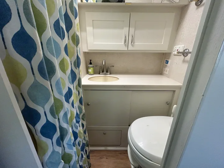 Compact RV rental bathroom features a vibrant blue/green patterned shower curtain, sink, toilet, & white storage cabinets.. Airstream Excella 2000