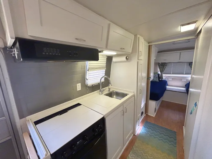 RV rental interior: A compact kitchen with gray backsplash, stove, sink, leading to a sleeping area with beds.. Airstream Excella 2000