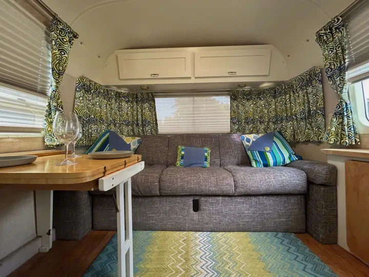 RV rental interior with cozy gray couch, table, colorful patterned curtains, and a bright rug creating a welcoming space.. Airstream Excella 2000