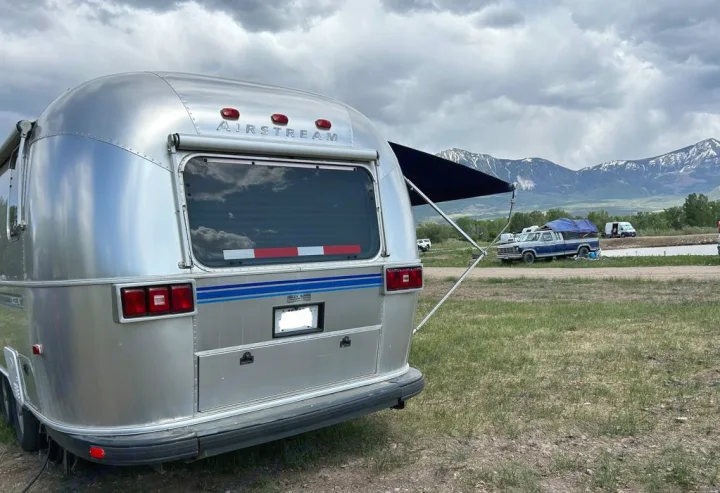 This Airstream rv rental, with its awning extended, offers stunning mountain views under a cloudy sky, ideal for camping.. Airstream Excella 2000