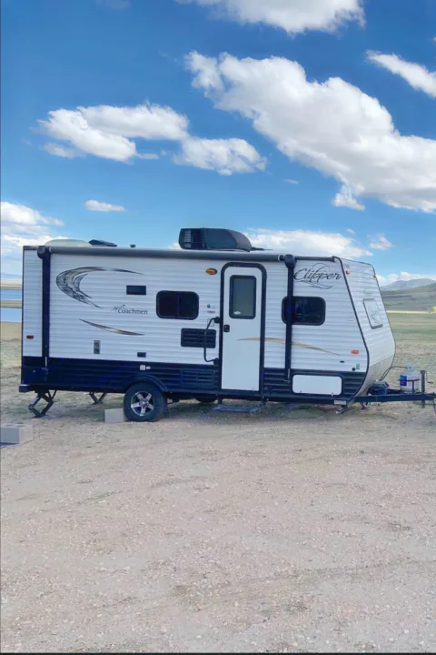 A white Coachmen Clipper travel trailer, ideal for an RV rental, parked on gravel near a scenic lake and mountains.. Coachmen Clipper 2015