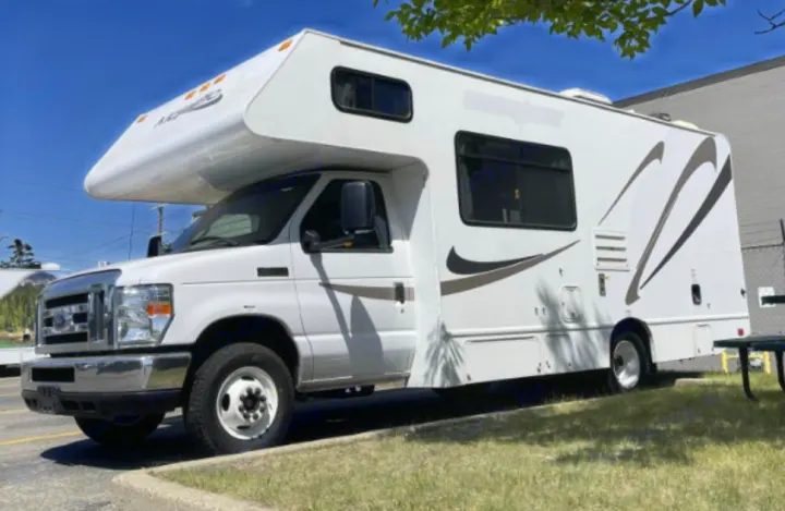 White Class C motorhome RV rental with brown decals, parked on grass & pavement on a sunny day.. Thor Majestic 2018