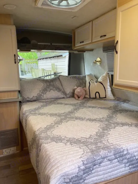Explore this cozy RV rental bedroom, featuring a patterned bed, cat pillow, and built-in cabinets, ready for your next adventure.. Winnebago Rialta 1998