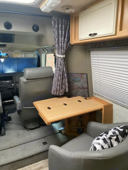 Inside an RV rental: a compact camper van interior with swiveling seats, a fold-down table, and patterned curtains.. Winnebago Rialta 1998