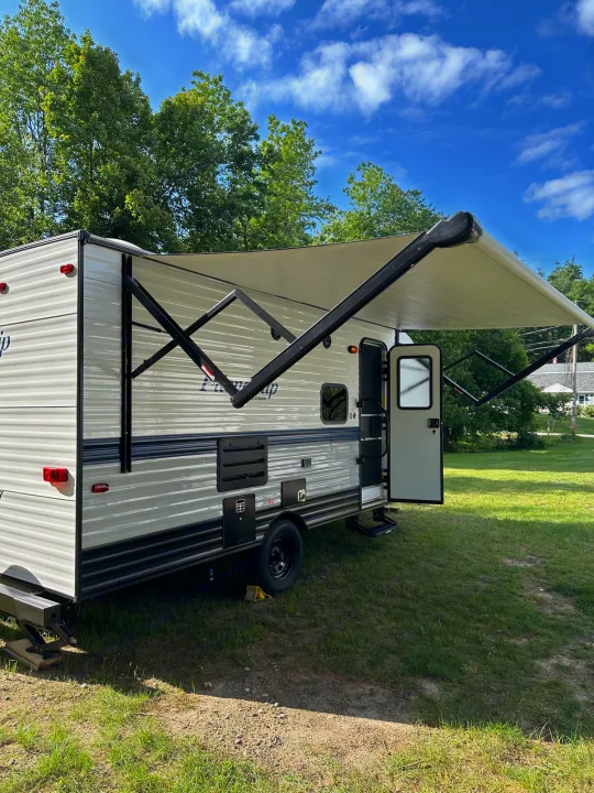 This RV rental, a light-colored travel trailer, is parked on grass with its awning extended and door open on a sunny day.. Gulf Stream Friendship 2022