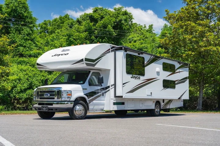 White Jayco Class C RV rental parked on asphalt, surrounded by green trees under a blue sky, ready for adventure.. Jayco Redhawk 2019