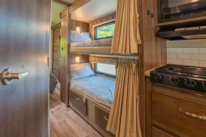 RV rental interior featuring cozy bunk beds with curtains, windows, and lamps, plus a compact kitchen with stovetop and microwave.. Jayco Redhawk 2019