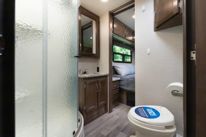 RV rental interior: a compact bathroom with a textured glass shower, toilet, and sink area, adjacent to the bedroom.. Jayco Redhawk 2019