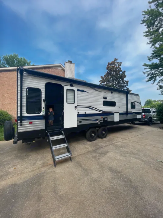 A young child peeks from a white and blue travel trailer, an RV rental, parked on a driveway with a pickup truck.. Keystone RV Springdale 2023
