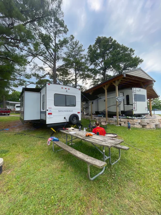 Two RVs, including a Springdale RV rental, are parked at a scenic campsite with a picnic area, shaded by trees, near a lake.. Keystone RV Springdale 2023