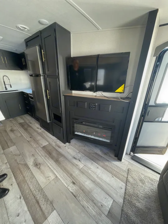 RV rental interior: Dark kitchen, stainless fridge, wood floors, and living area with TV & electric fireplace.. Keystone RV Springdale 2023