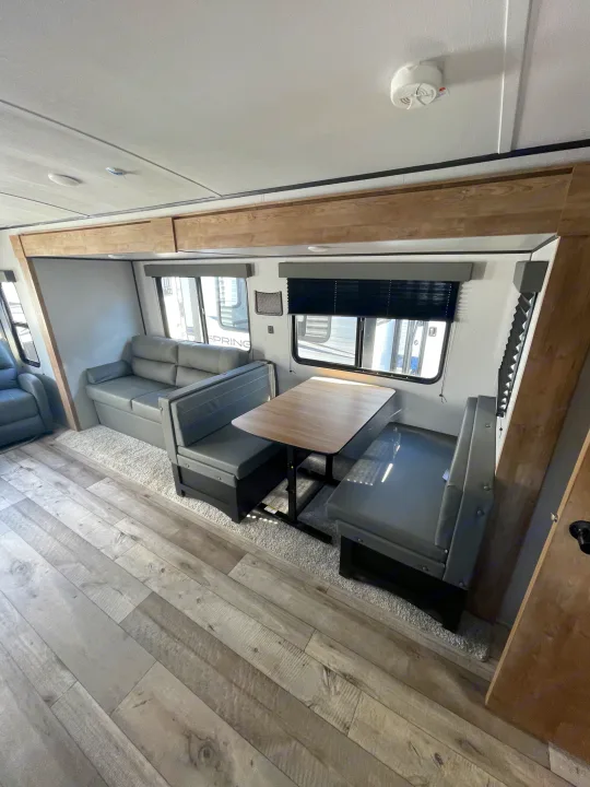 Bright RV rental interior with grey sofa, dining booth, wooden table, and light wood-look flooring.. Keystone RV Springdale 2023