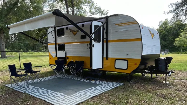 A vibrant yellow and white travel trailer RV rental with an awning extended over a mat and four folding chairs, perfect for camping.. Riverside RV Retro 2022