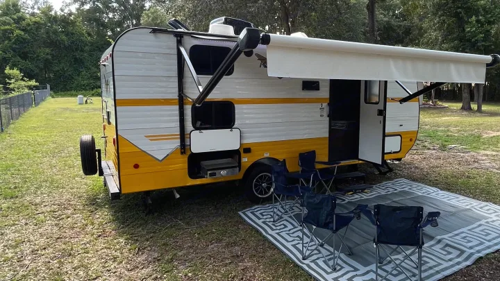 RV rental setup: White & yellow travel trailer with deployed awning, grill, & four camp chairs on a mat, ready for outdoor fun.. Riverside RV Retro 2022
