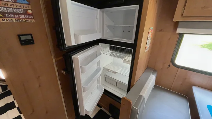 Inside an RV rental: An empty, compact refrigerator and freezer with doors open, revealing white shelves, ready for stocking.. Riverside RV Retro 2022