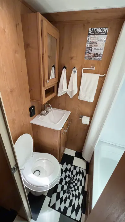 This cozy RV rental bathroom boasts wood walls, white fixtures, and a quirky rules sign above its checkered floor.. Riverside RV Retro 2022
