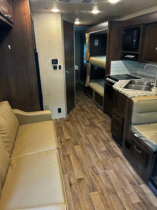 RV rental interior: light sofa, wood flooring, compact kitchen with stove/sink, and built-in bunk beds.. Jayco Redhawk 2020