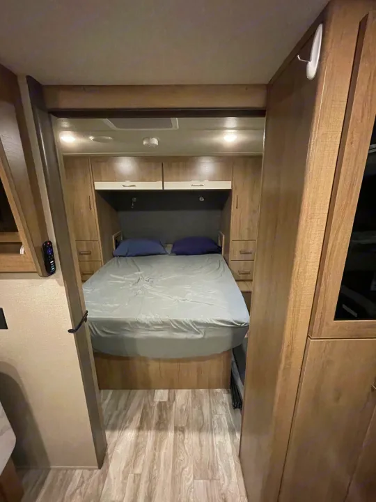 Cozy RV rental bedroom featuring a comfortable bed with blue pillows, ample storage, and stylish wood-grain interior.. Grand Design Imagine XLS 2022
