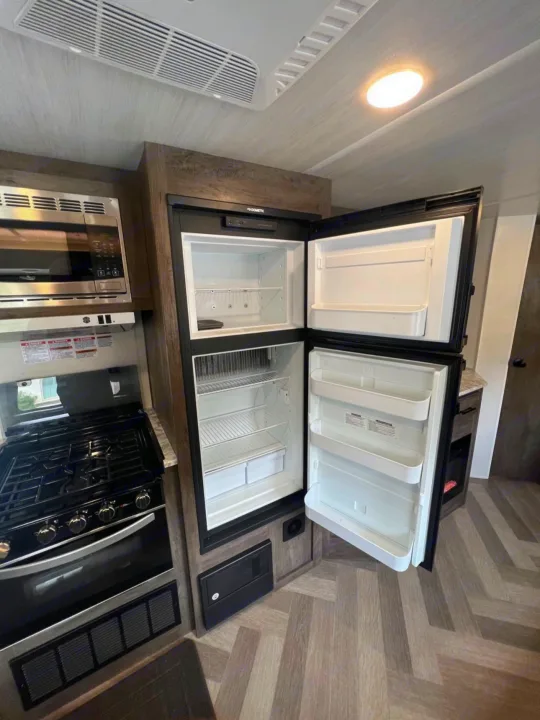 RV rental kitchen: View of open Dometic fridge, microwave, gas stove, and oven. Features ceiling AC & modern interior.. Forest River Wildwood 2021