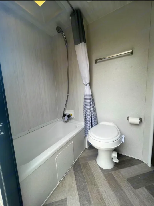 An RV rental's modern bathroom features a compact bathtub with shower, a toilet, and gray wood-look finishes.. Forest River Wildwood 2021