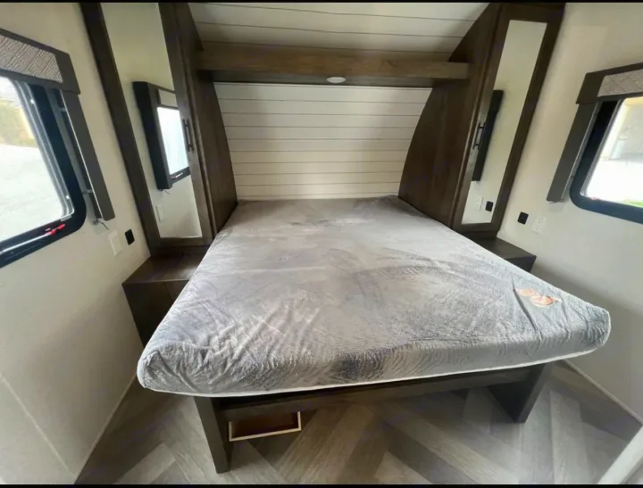 Step into this modern RV rental bedroom: comfy bed, integrated nightstands with mirrors, overhead storage, and windows.. Forest River Wildwood 2021