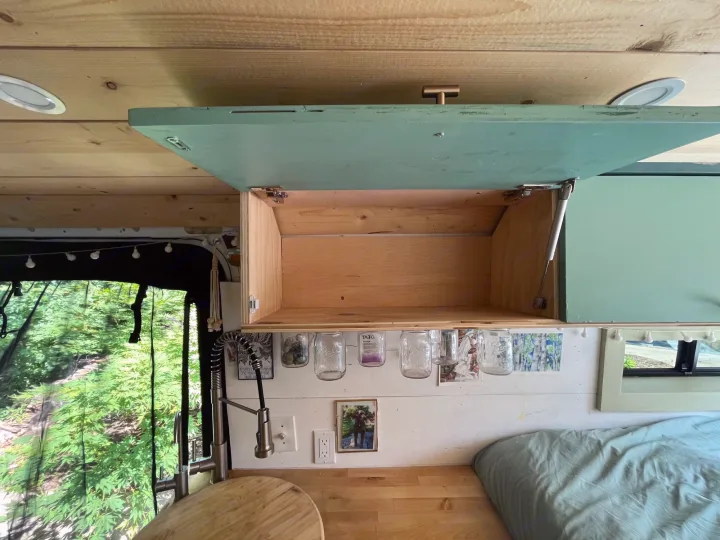 Cozy RV rental interior with overhead storage, a compact kitchen, and a bed. Green tree view outside.. Other Sprinter Van 2017