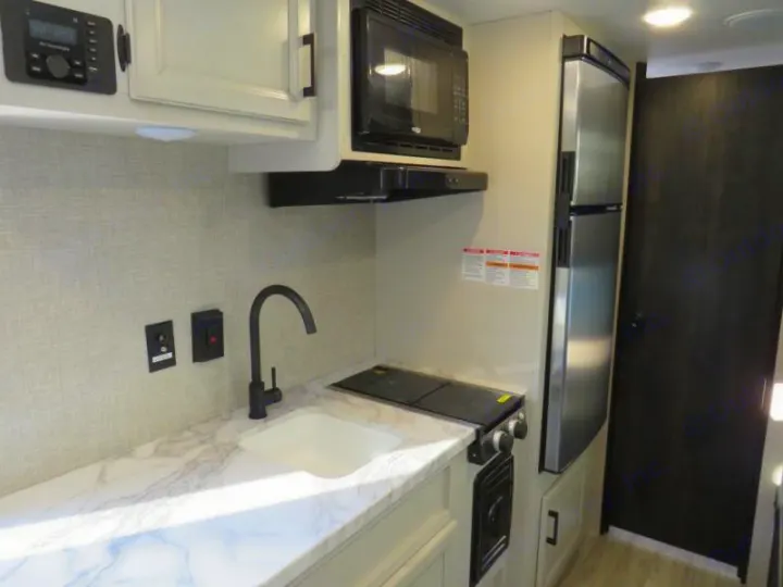 Clean, modern RV rental kitchen area with a white sink, black faucet, stovetop, microwave, and compact refrigerator.. Jayco Jay Flight SLX STX Edition 2023
