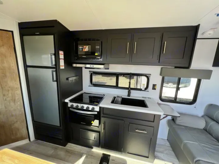 Modern RV rental kitchen interior: large fridge, stove, microwave, sink, dark cabinets, and adjacent sofa.. Keystone RV Springdale 2022
