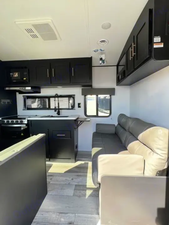 Modern RV rental interior: black kitchen cabinets with stove/sink on left, gray sofa/living area on right.. Keystone RV Springdale 2022