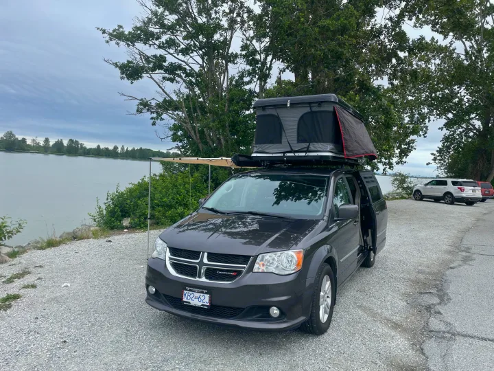 Enjoy nature with this RV rental: a Dodge minivan featuring a rooftop tent & awning, parked by a lake. Perfect for camping!. Dodge Caravan 2015