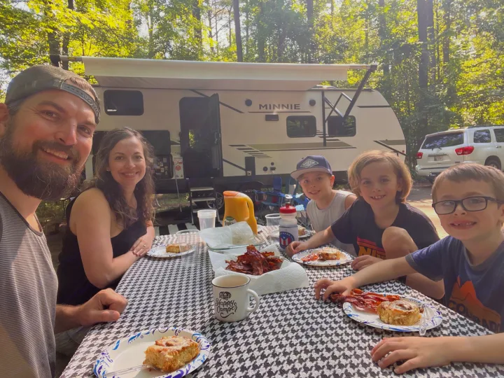 Smiling family enjoys an outdoor breakfast by their Minnie RV rental at a sunny campground.. Winnebago Minnie 2022
