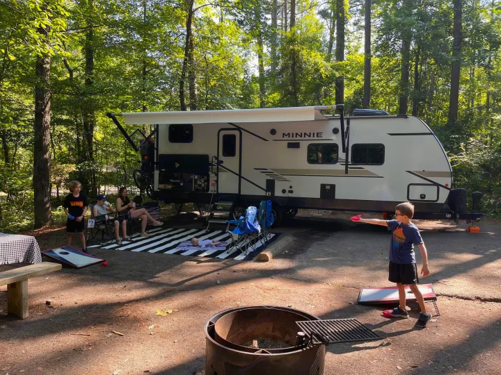 Family camping with an RV rental. Kids play cornhole while adults relax at a sunny, wooded campsite.. Winnebago Minnie 2022