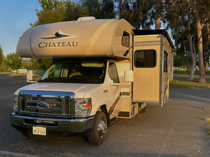 This light-colored Chateau Class C RV rental is parked with its side extended, ready for a travel adventure among trees.. Thor Chateau 2020