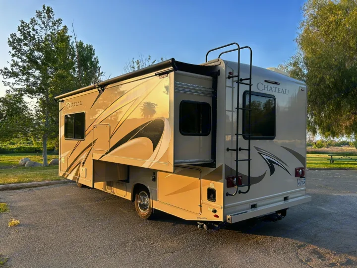 This Chateau Class C RV rental with an extended slide-out and awning is parked on asphalt, surrounded by trees.. Thor Chateau 2020