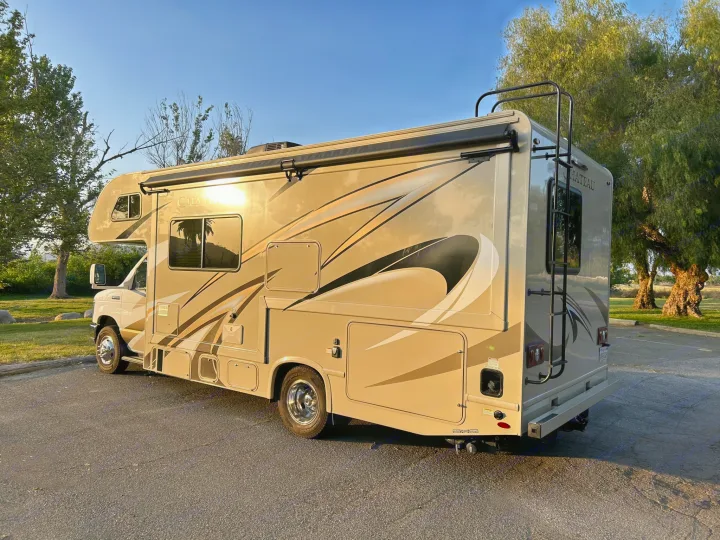 A tan Class C RV rental, featuring graphics and an awning, is parked on asphalt with trees in the background.. Thor Chateau 2020