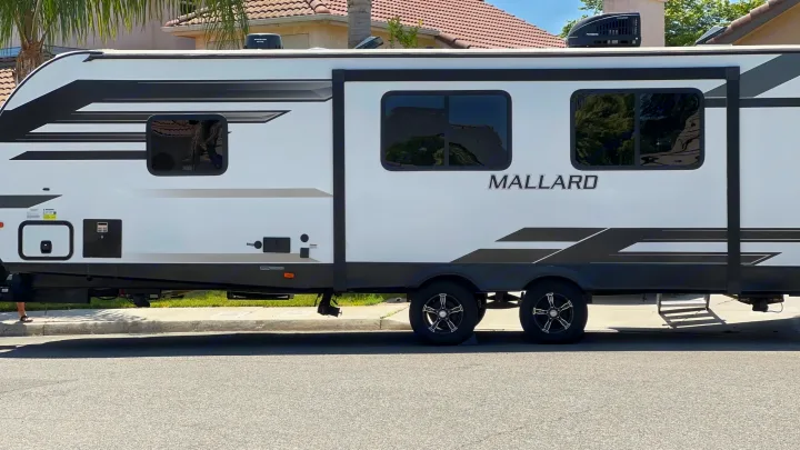 Side view of a white Mallard travel trailer RV rental with black graphics, windows, and dual axles, parked by a curb.. Heartland Other 2021