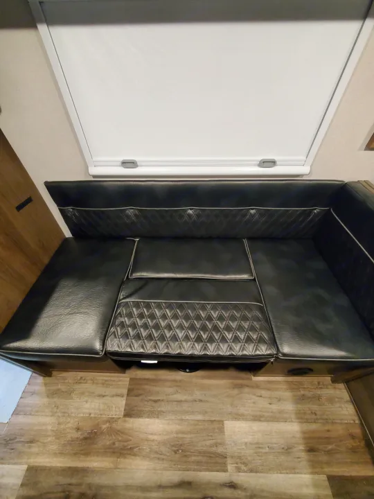 An L-shaped black faux leather RV dinette bench with a white window blind and wood-look flooring, great for any rv rental.. Imperial Outdoors XploreRV 2023