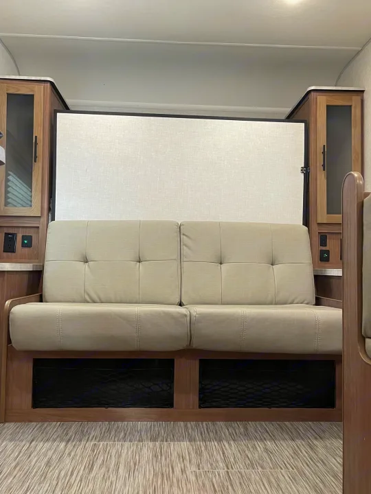 RV rental interior: a light sofa, under-sofa mesh storage, wood cabinets with outlets, and a murphy bed panel.. Forest River No Boundary 2020
