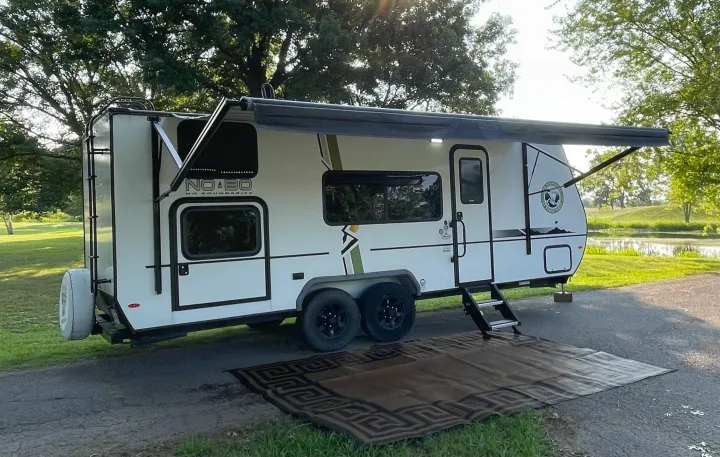 This travel trailer RV rental, with an extended awning and mat, is parked by a pond, ideal for outdoor adventures.. Forest River No Boundary 2020