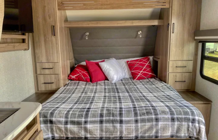 Cozy RV rental bedroom with a plaid blanket, red pillows, and wooden built-in storage. Features a TV and window.. Grand Design Other 2023