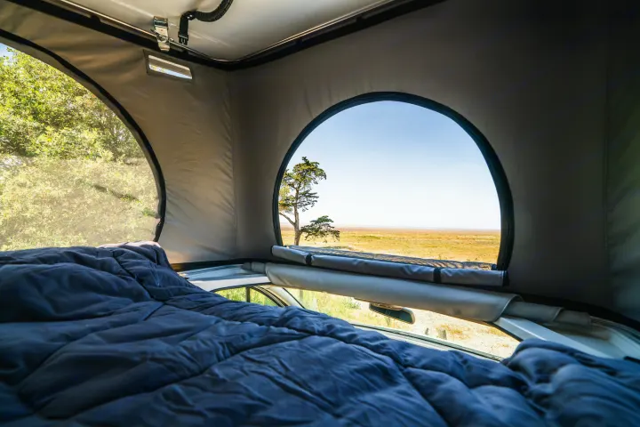 Cozy RV rental bed under pop-top windows, offering serene views of trees and a wide-open landscape.. HANROAD TREK 4 2023