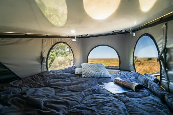 A plush bed inside an RV rental pop-top tent, offering panoramic window views of diverse outdoor landscapes. Ideal for camping.. HANROAD TREK 4 2023