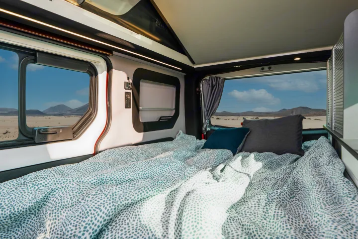 This RV rental features a cozy bed with patterned sheets, offering panoramic desert mountain views from its large windows.. HANROAD TREK 4 2023