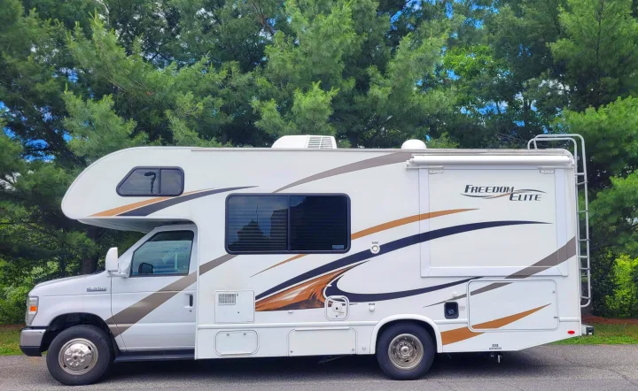 White RV rental, side view, parked outdoors with trees. Features "Freedom Elite" graphics, awning, and roof ladder.. Thor Freedom Elite 2017