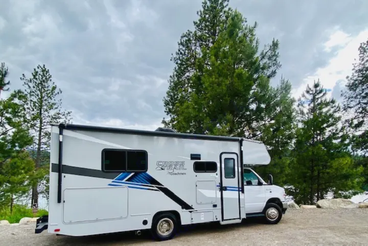 Experience nature with this RV rental: a modern motorhome parked by tall pine trees & a serene lake under a cloudy sky.. Forest River Coachmen Cross Trail XL 2022