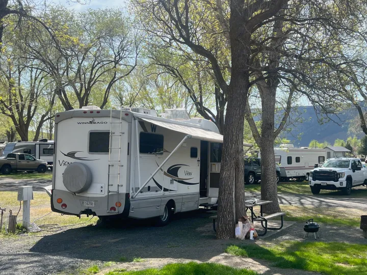 White Winnebago View RV rental at a green campground with trees. Awning extended, picnic table, grill, and hookups visible.. Winnebago View 2008