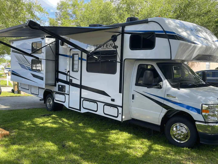 This white and blue Forest River Forester RV rental, featuring an extended awning and slide-out, is parked on green grass.. Forester LE 2023
