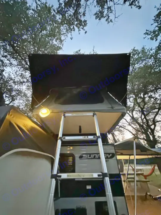 RV rental travel trailer with rooftop tent, illuminated by an outdoor light, and ladder for easy camping access.. Sunset Park RV SunRay 2023