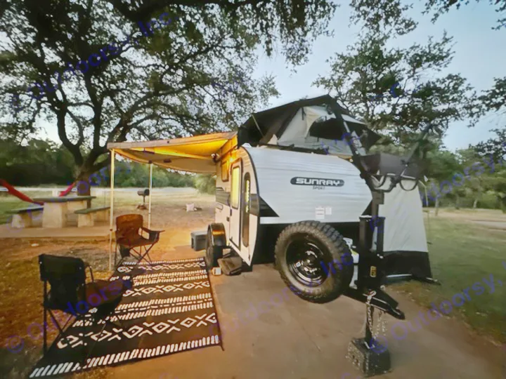 Nighttime rv rental setup: a small teardrop trailer with illuminated awning, camping chairs, and a rooftop tent. Perfect for adventure.. Sunset Park RV SunRay 2023